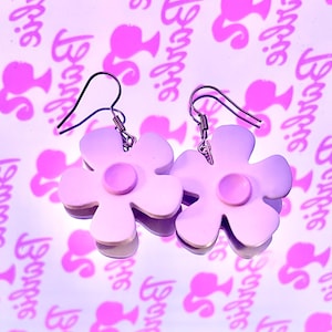 Puede incluir: Un par de pendientes colgantes en forma de flor lavanda con un pequeño círculo rosa en el centro de cada flor. Los pendientes tienen un gancho plateado.