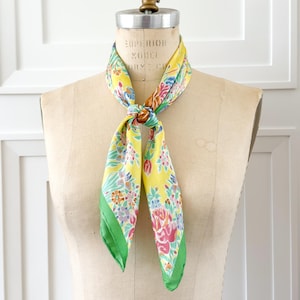 May include: A yellow silk scarf with a floral pattern in pink, blue, and orange. The scarf has a green border and is tied in a knot. The scarf is displayed on a vintage dress form.