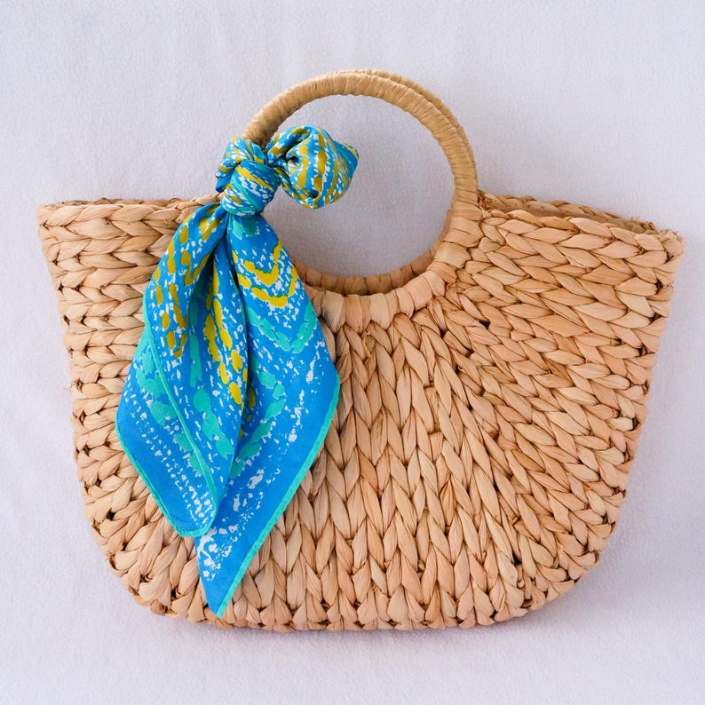 May include: Woven straw tote bag with a circular handle and a blue and yellow patterned scarf tied to the handle. The bag is a light tan color and has a wide, open top.
