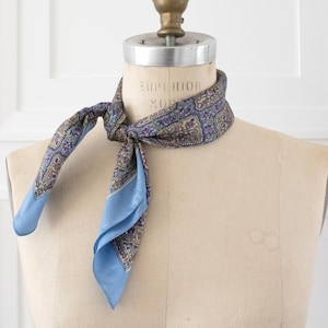 May include: A square silk scarf tied around the neck of a mannequin. The scarf features a complex paisley and floral pattern in shades of blue, purple, and gold, with a solid light blue border along the edges of the fabric.