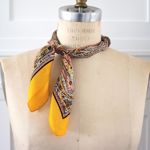 May include: A vintage silk scarf with a vibrant paisley pattern and bright yellow borders. The scarf is knotted around a mannequin's neck, showcasing its colorful design with shades of red, pink, and green. The scarf's edges are outlined in black.