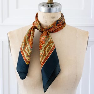 May include: A square scarf tied around a mannequin neck, featuring a dark navy blue border and a central pattern of intricate floral and paisley motifs in shades of burnt orange, gold, cream, and muted green, with a smooth, satin-like finish.