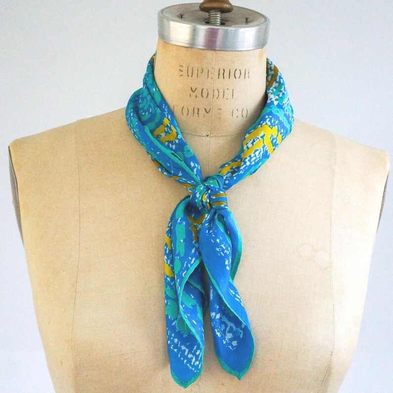 May include: A vintage blue scarf with a vibrant pattern of yellow and teal designs. The scarf is tied in a knot, showcasing its flowing fabric and intricate details. The scarf is displayed on a mannequin.