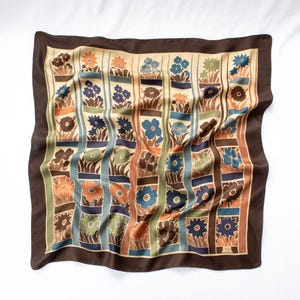 May include: A square scarf featuring a grid pattern of floral illustrations in shades of blue, orange, green, and brown. The design is framed by a solid dark brown border, with each rectangular section containing stylized flowers and foliage.