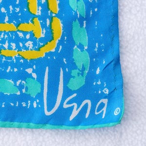 May include: Close-up of a square, turquoise fabric with a white border. The fabric features yellow and teal abstract shapes and the word "Usna" in white script.
