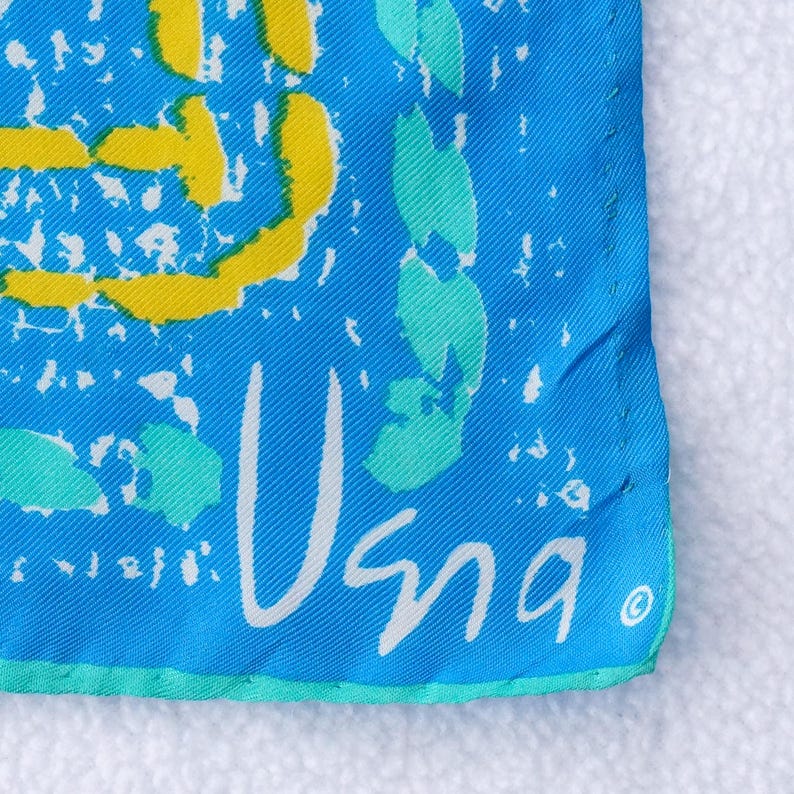 May include: Close-up of a square, turquoise fabric with a white border. The fabric features yellow and teal abstract shapes and the word "Usna" in white script.