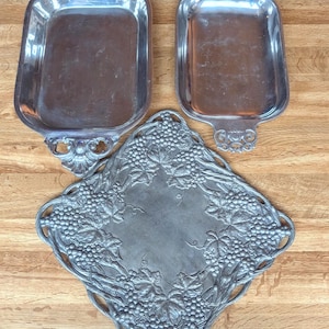 May include: Three silver-toned serving trays. Two are rectangular with ornate handles. The third is a diamond shape, featuring a grape and leaf design around the edges. All are on a wooden surface.