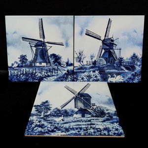 May include: Three blue and white ceramic tiles depicting Dutch windmills in a landscape. The tiles feature detailed illustrations of windmills, trees, water, and clouds. The artwork is in a traditional Delftware style.