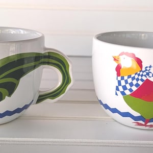 May include: Two white ceramic mugs with a colorful rooster design. The rooster features red, yellow, and green accents, a blue and white checkered scarf, and a blue wave pattern at the base. The handles are green.