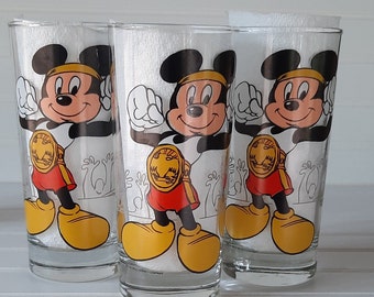 May include: Three clear glass cups with a cartoon image of Mickey Mouse as a wrestler. Each cup features a different pose of Mickey Mouse wearing a red and yellow wrestling outfit. The cups are printed with the text "The Walt Disney Company".