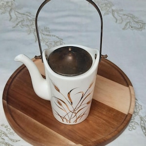 May include: A white ceramic teapot with a wheat design in gold, featuring a metal handle and lid. The teapot is placed on a round wooden tray with a two-tone design. The spout is on the left side.