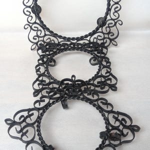 May include: Black metal decorative candle holder with three circular frames connected by ornate, scrolled metalwork. The frames have a twisted rope-like border and are embellished with decorative flourishes.