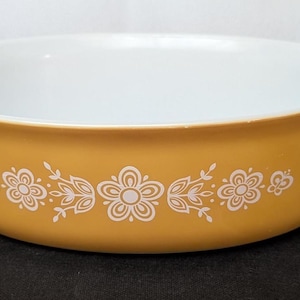 May include: A round, shallow casserole dish with integrated side handles. The exterior is a solid mustard yellow color, featuring a white floral pattern around the center. The interior of the dish is smooth, glossy white, and the rim is wide.