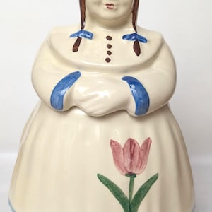 May include: A ceramic cookie jar shaped like a person wearing a cream-colored dress with a pink tulip design, a blue bonnet, and blue sleeve cuffs. The figure has brown hair in braids with blue bows and a painted face with blue eyes and red lips.