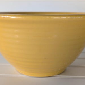 May include: A yellow ceramic bowl with a wide, rounded shape. The bowl has a smooth, glossy finish and a slightly flared rim. The exterior features subtle, horizontal ridges. This bowl is suitable for serving or display.