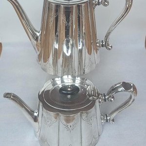 May include: A tall silver coffee pot and a shorter silver teapot, both featuring a fluted body design, ornate curved handles, and black circular finials on their lids. The silver surfaces are decorated with delicate engraved patterns.