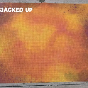 May include: A cross-stitch canvas with a gradient of orange, yellow, and purple hues. The words "JACKED UP" are printed in white at the top left corner. The canvas has a textured surface, with a grid pattern.