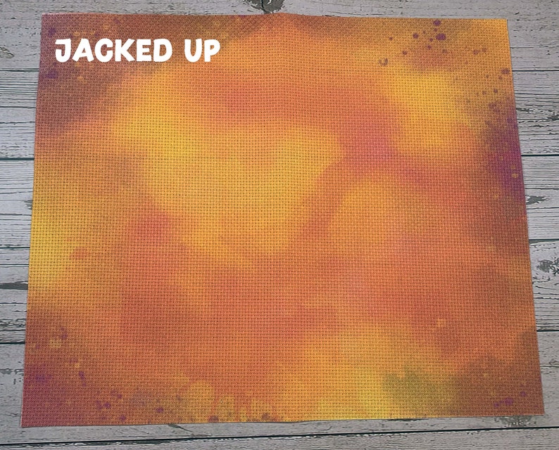 May include: A cross-stitch canvas with a gradient of orange, yellow, and purple hues. The words "JACKED UP" are printed in white at the top left corner. The canvas has a textured surface, with a grid pattern.