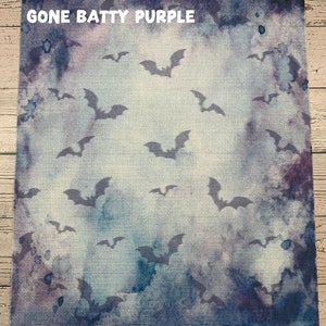 May include: A square fabric with a watercolor-style background in shades of blue and purple. Silhouetted bats are scattered across the surface. The text "GONE BATTY PURPLE" is at the top.