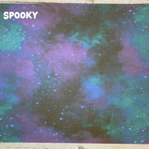 May include: A rectangular fabric with a cosmic design. The background is a dark, textured canvas with a nebula-like pattern in shades of purple, teal, and blue. The word "SPOOKY" is printed in white in the upper left corner.