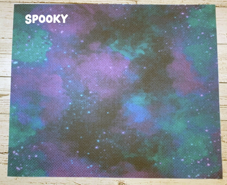 May include: A rectangular fabric with a cosmic design. The background is a dark, textured canvas with a nebula-like pattern in shades of purple, teal, and blue. The word "SPOOKY" is printed in white in the upper left corner.