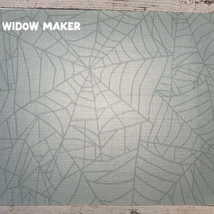 May include: A rectangular placemat with a light blue spiderweb design. The text "WIDOW MAKER" is printed in white in the upper left corner. The placemat has a textured appearance.
