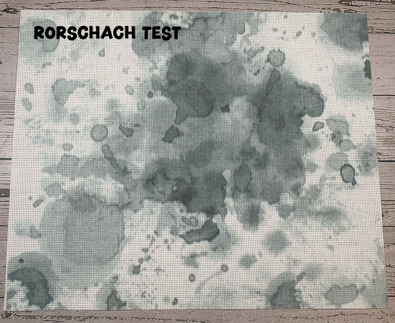 May include: A rectangular textile with a white background and a pattern of gray watercolor-like blotches and splatters. The text "RORSCHACH TEST" is printed in black at the top. The textile has a cross-stitch appearance.