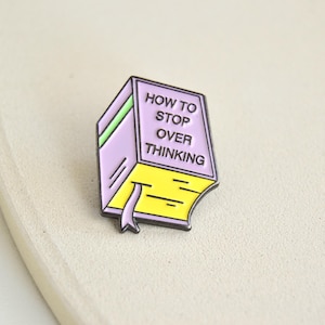 Könnte beinhalten: Ein farbenfroher Emaille-Pin in Form eines Buches. Der Pin hat einen lila Einband mit den Worten "HOW TO STOP OVER THINKING" in Weiß. Die Seiten sind gelb, und ein lila Lesezeichen ist sichtbar.