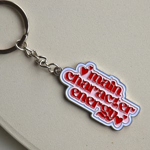 May include: A silver keychain with a rectangular charm. The charm has a white border, a light blue outline, and the words "main character energy" in red, with small red heart accents. The keychain is on a light beige surface.