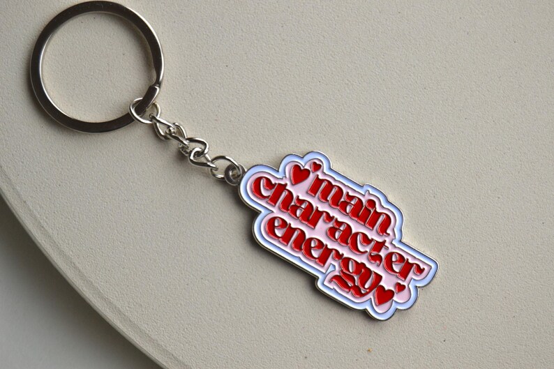 May include: A silver keychain with a rectangular charm. The charm has a white border, a light blue outline, and the words "main character energy" in red, with small red heart accents. The keychain is on a light beige surface.
