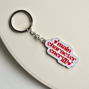 May include: A silver keychain with a rectangular charm. The charm has the words "main character energy" in red, outlined in white and blue, with red heart accents. The keychain is on a light beige surface.