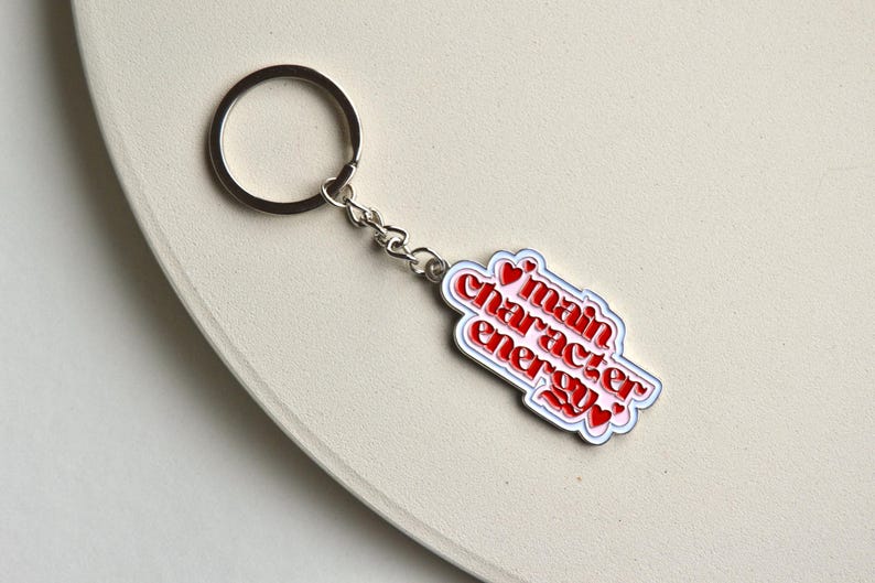May include: A silver keychain with a rectangular charm. The charm has the words "main character energy" in red, outlined in white and blue, with red heart accents. The keychain is on a light beige surface.