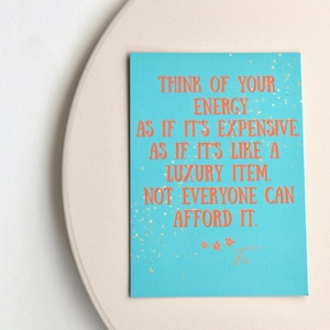 Könnte beinhalten: Türkisfarbene Karte mit orangefarbenem Text: "DENKE AN DEINE ENERGIE, ALS WÄRE SIE TEUER, ALS WÄRE SIE EIN LUXUSARTIKEL. NICHT JEDER KANN ES SICH LEISTEN." Goldene Flecken und eine cremefarbene Oberfläche.