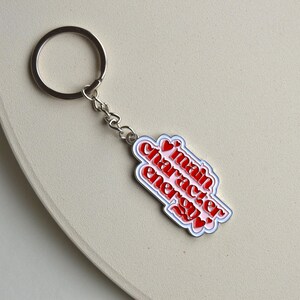 May include: A silver keychain with a charm that reads "main character energy" in red text with white and red accents. The keychain is on a light beige surface.