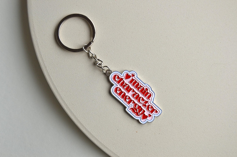May include: A silver keychain with a charm that reads "main character energy" in red text with white and red accents. The keychain is on a light beige surface.