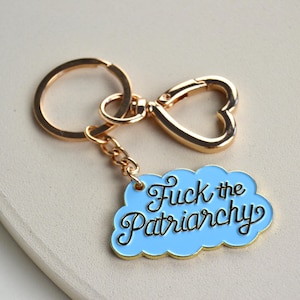 Könnte beinhalten: Ein goldfarbener Schlüsselanhänger mit herzförmigem Verschluss und kleiner Kette. Der Anhänger hat einen hellblauen, wolkenförmigen Anhänger mit dem Schriftzug "Fuck the Patriarchy" in schwarzer Schrift.