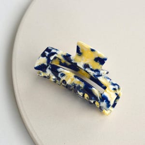 Könnte beinhalten: Eine rechteckige Haarspange mit einem marmorierten Muster in Gelb- und Marineblautönen. Die Spange besteht aus glänzendem Material und hat einen festen Halt. Dieses Haaraccessoire ist perfekt für verschiedene Frisuren.