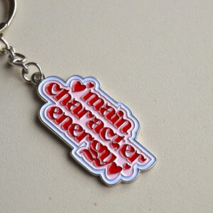 May include: A keychain with a silver chain and ring. The pendant is a white, pink, and red design with the words "main character energy" in a stylized font. Small red hearts are also included.