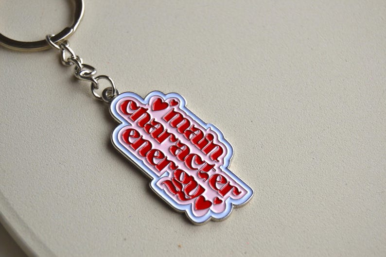 May include: A keychain with a silver chain and ring. The pendant is a white, pink, and red design with the words "main character energy" in a stylized font. Small red hearts are also included.