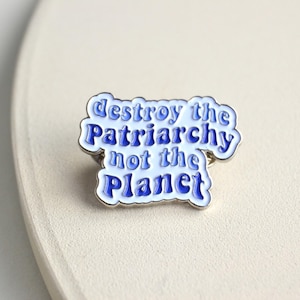 Könnte beinhalten: Blau-weißer Emaille-Pin mit dem Text "destroy the Patriarchy not the Planet". Der Pin hat einen silbernen Rand und einen Pin-Rücken. Der Text ist in einer stilisierten Schriftart.
