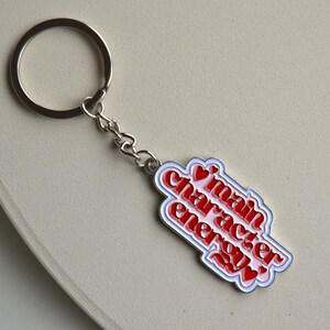 May include: A silver keychain with a rectangular charm. The charm has the words "main character energy" in red, outlined in white and blue, with red heart accents. The keychain is on a light beige surface.