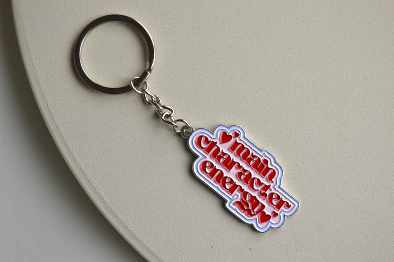 May include: A silver keychain with a rectangular charm. The charm has the words "main character energy" in red, outlined in white and blue, with red heart accents. The keychain is on a light beige surface.