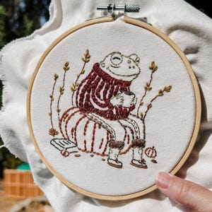 May include: Embroidered artwork in a wooden hoop. The design features a smiling frog wearing a striped sweater and boots, holding a mug. The frog is seated on a pumpkin, with sprigs of foliage around it. The embroidery is in shades of brown, green, and red.