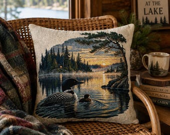 Può includere: Un cuscino quadrato con un design a arazzo intrecciato che raffigura una strolaga con il suo piccolo che nuotano su un lago al tramonto. La scena include una fitta foresta, una riva rocciosa con un pino e luce dorata riflessa sull'acqua.