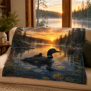 May include: A rectangular throw blanket featuring a printed scene of a loon with a chick on its back swimming in a lake at sunset. The image displays a golden sun over a forest, calm water with lily pads, and a small cabin on the distant shore.