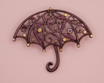 May include: An umbrella-shaped brooch with a dark bronze frame and purple accents. The brooch features a decorative openwork design with clear and iridescent crystals. The handle is curved, and the overall design is ornate.