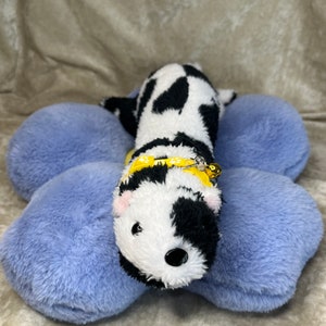 May include: A black and white spotted plush toy with a yellow collar and a bell. The toy is lying on a blue plush flower-shaped pillow.