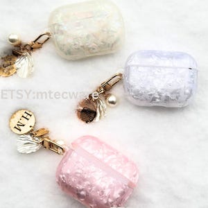 May include: Three clear Airpod cases with a sparkly, iridescent finish. Each case has a gold chain with a charm attached. The charms include a pearl, a small shell, and a gold disc with the word "Faith" on it. The cases are available in white, pink, and iridescent.
