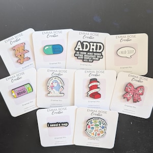 Può includere: Una collezione di spille smaltate su cartoncini bianchi. Le spille presentano vari disegni, tra cui una pillola, un fiocco e testi che recitano "ADHD", "I need sleep" e "I need a nap". Le spille sono colorate e hanno una finitura lucida.