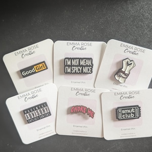 Puede incluir: Una colección de pines de esmalte sobre tarjetas blancas. Los pines presentan varios diseños, incluyendo textos que dicen "Good Girl", "I'M NOT MEAN, I'M SPICY NICE", "PARENTAL ADVISORY EXPLICIT CONTENT", "CHOKE ME" y "smut club". Los pines son negros, blancos y rosas.